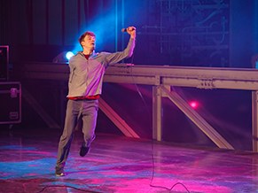 Comedian performing on a lit stage set with the mic held high