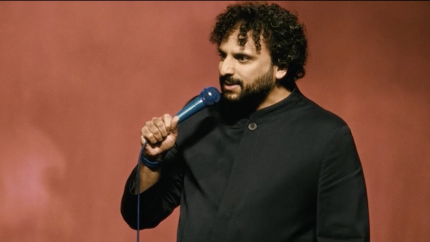 Nish Kumar on stage with a blue microphone against a red backdrop