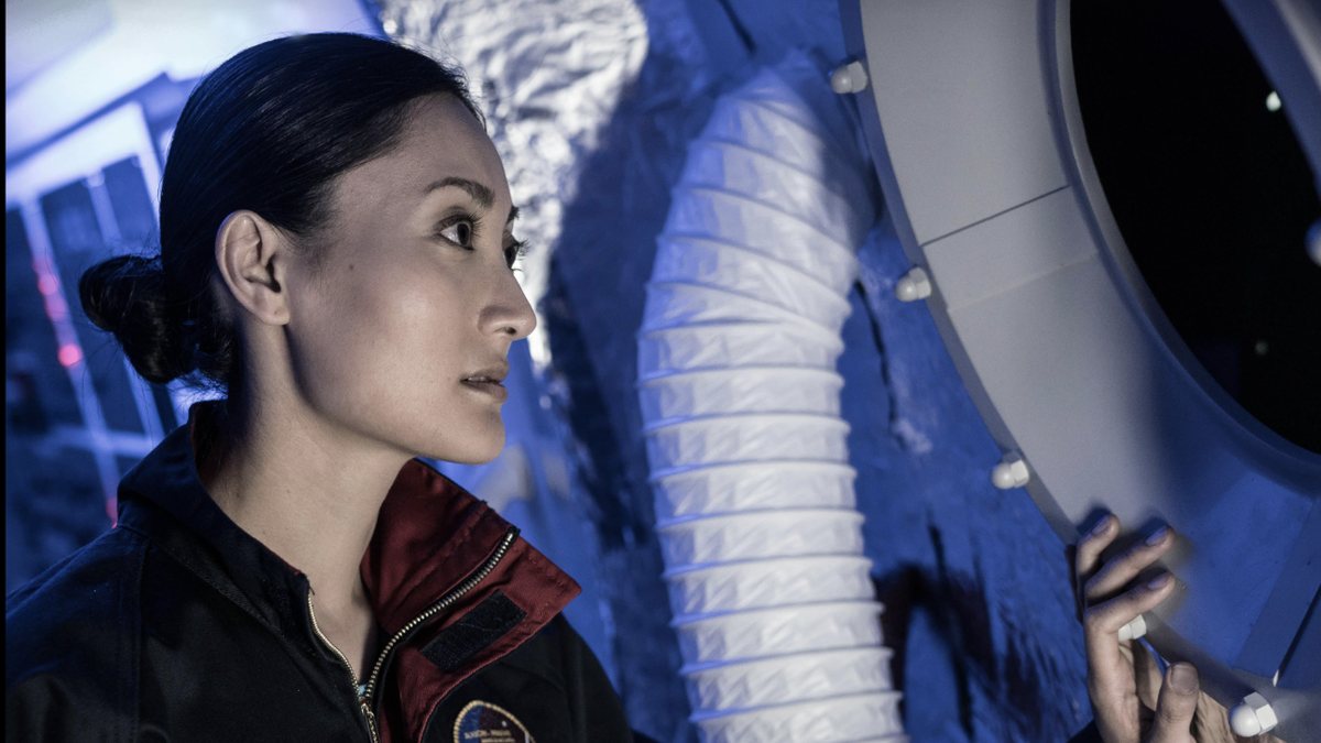 An astronaut in a flight suit looking out of a spacecraft porthole, from the This Is Axiom poster